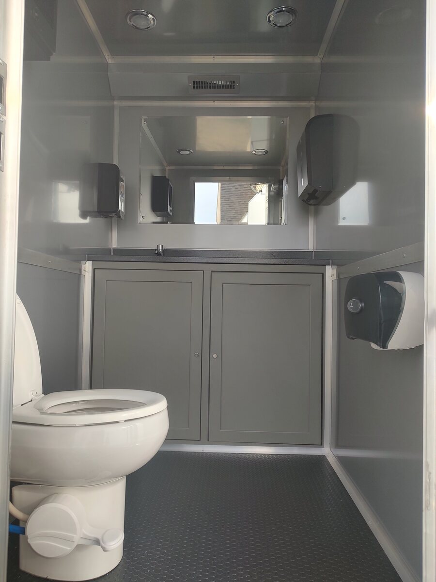 Inside a restroom trailer: a stall with a flushing toilet, a vanity and bright lighting