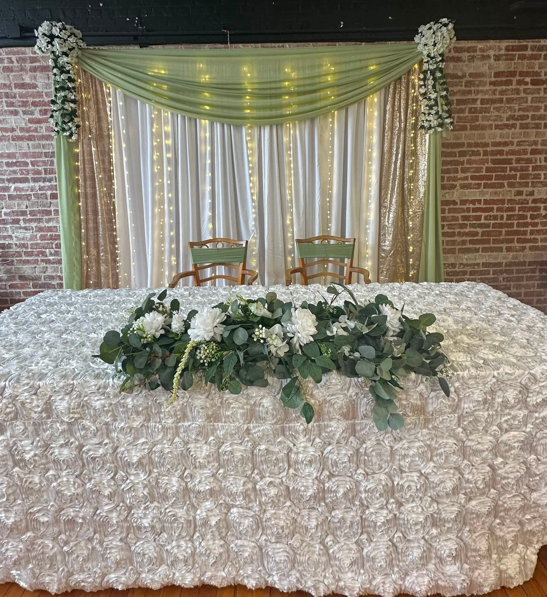 Sweetheart table with a rosette linen and eucalyptus and white floral garland, in front of a draped backdrop with string lights at Pleasant Street Hall